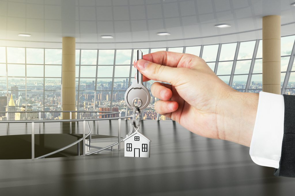 Hand übergibt Wohnungsschlüssel mit Hausanhänger vor Panoramafenster mit Stadtblick