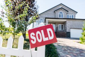 Rotes "SOLD"-Schild hängt an einem weißen Lattenzaun vor einem verkauften Einfamilienhaus aus hellem Backstein