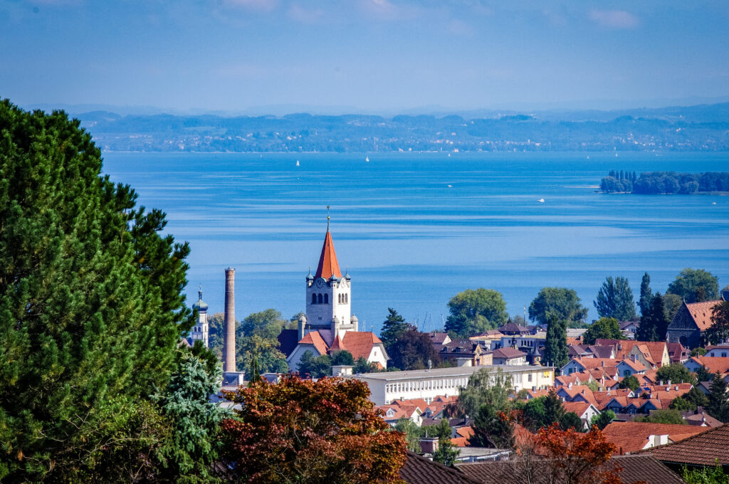 Blick über Dächer und einen Kirchturm mit rotem Spitzdach auf den Bodensee mit Segelbooten und Hügeln am Horizont