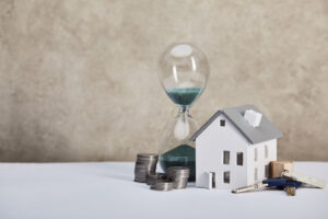 Sanduhr, Münzstapel und Hausmodell mit Schlüsselbund symbolisieren Zeit und Kosten beim Immobilienverkauf