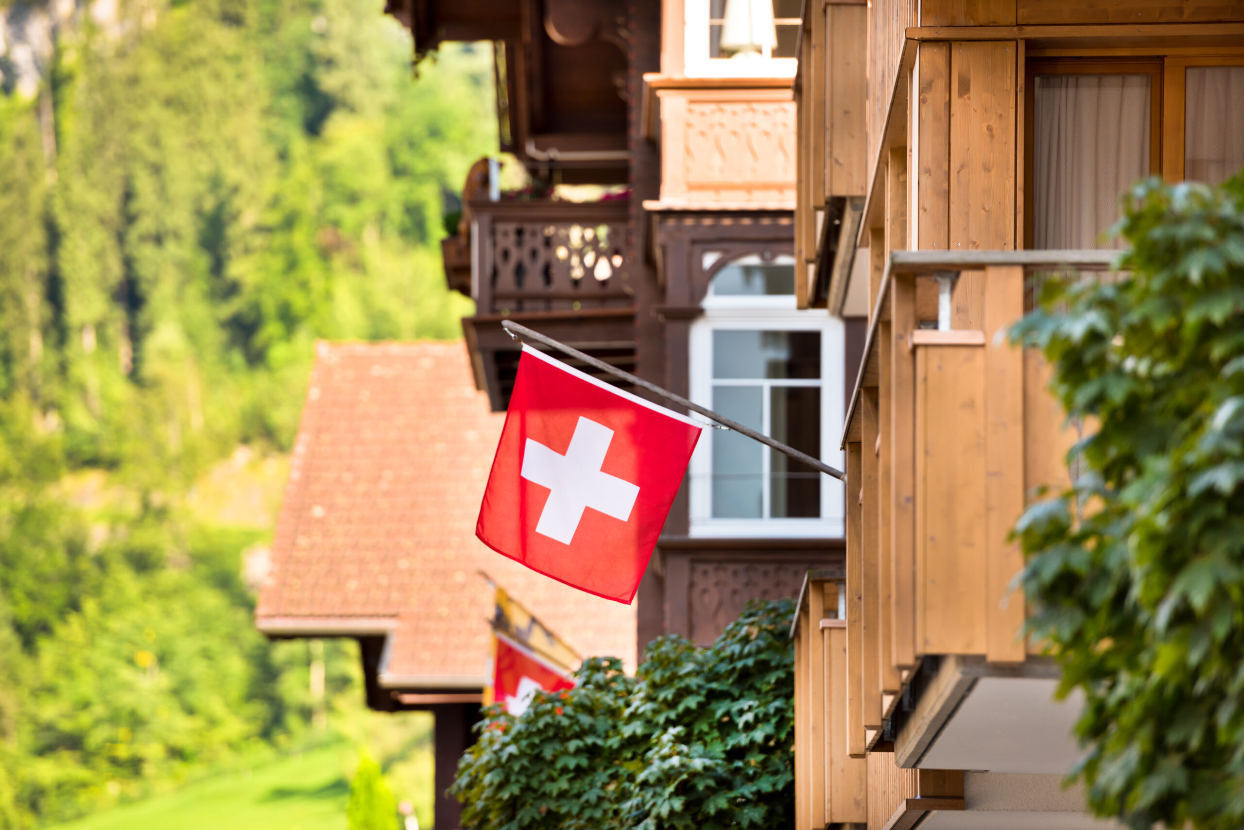 Schweizer Flagge weht an einem traditionellen Holzchalet mit Balkon, im Hintergrund bewaldete Berghänge